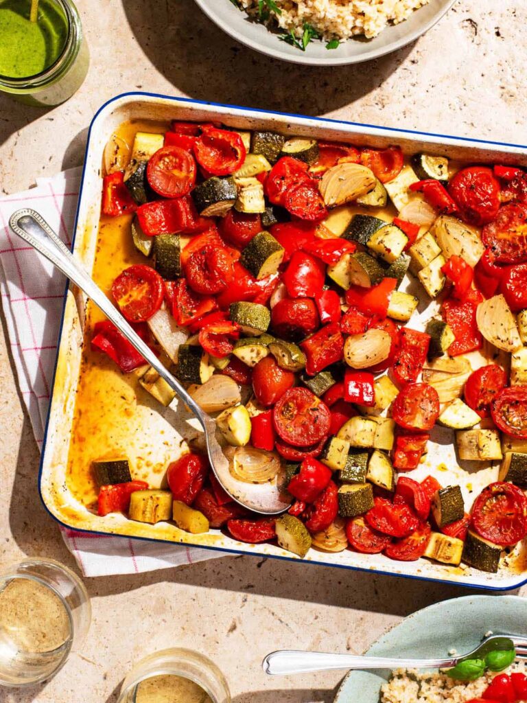 Roasted summer vegetables on a baking sheet with bowls and glasses around.