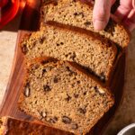 Banana bread with walnuts on a cutting board, a woman's hand taking a slice.