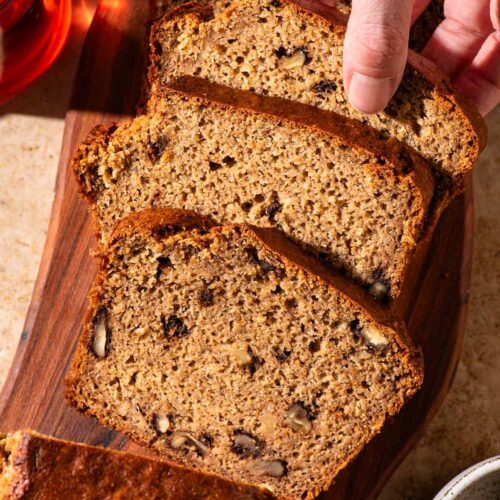Banana bread with walnuts on a cutting board, a woman's hand taking a slice.