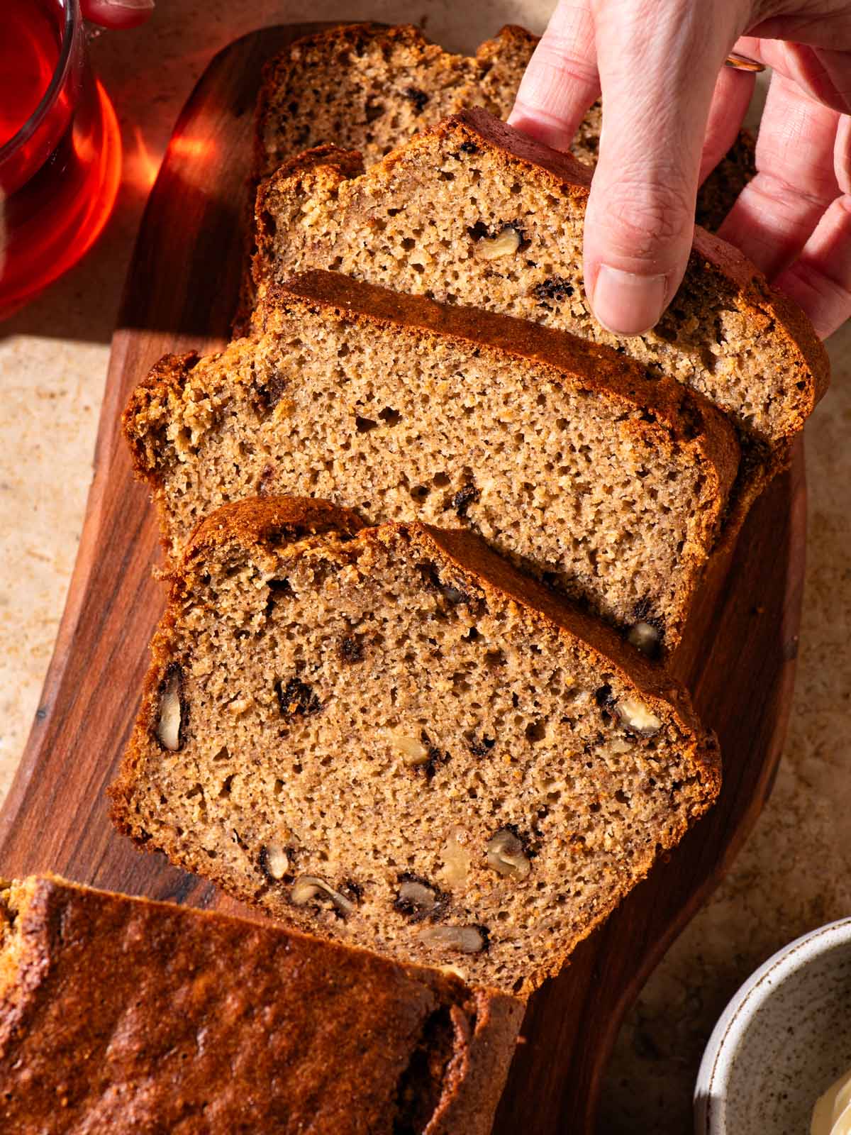 Banana bread with walnuts on a cutting board, a woman's hand taking a slice.
