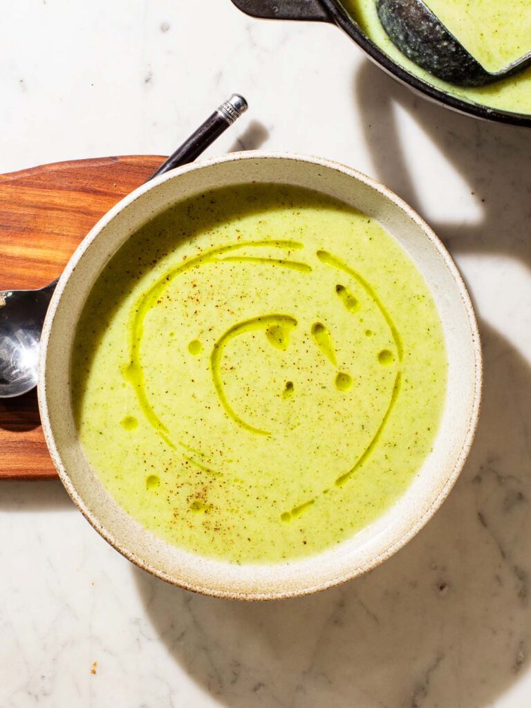 A bowl of light green soup on a wooden board with a spoon.