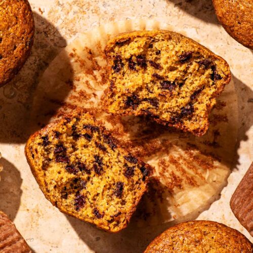 Several chocolate chip muffins on a stone surface, one halved to show interior texture.