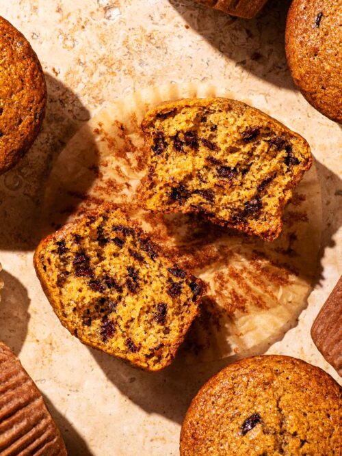 Several chocolate chip muffins on a stone surface, one halved to show interior texture.