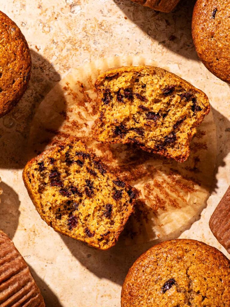 Several chocolate chip muffins on a stone surface, one halved to show interior texture.